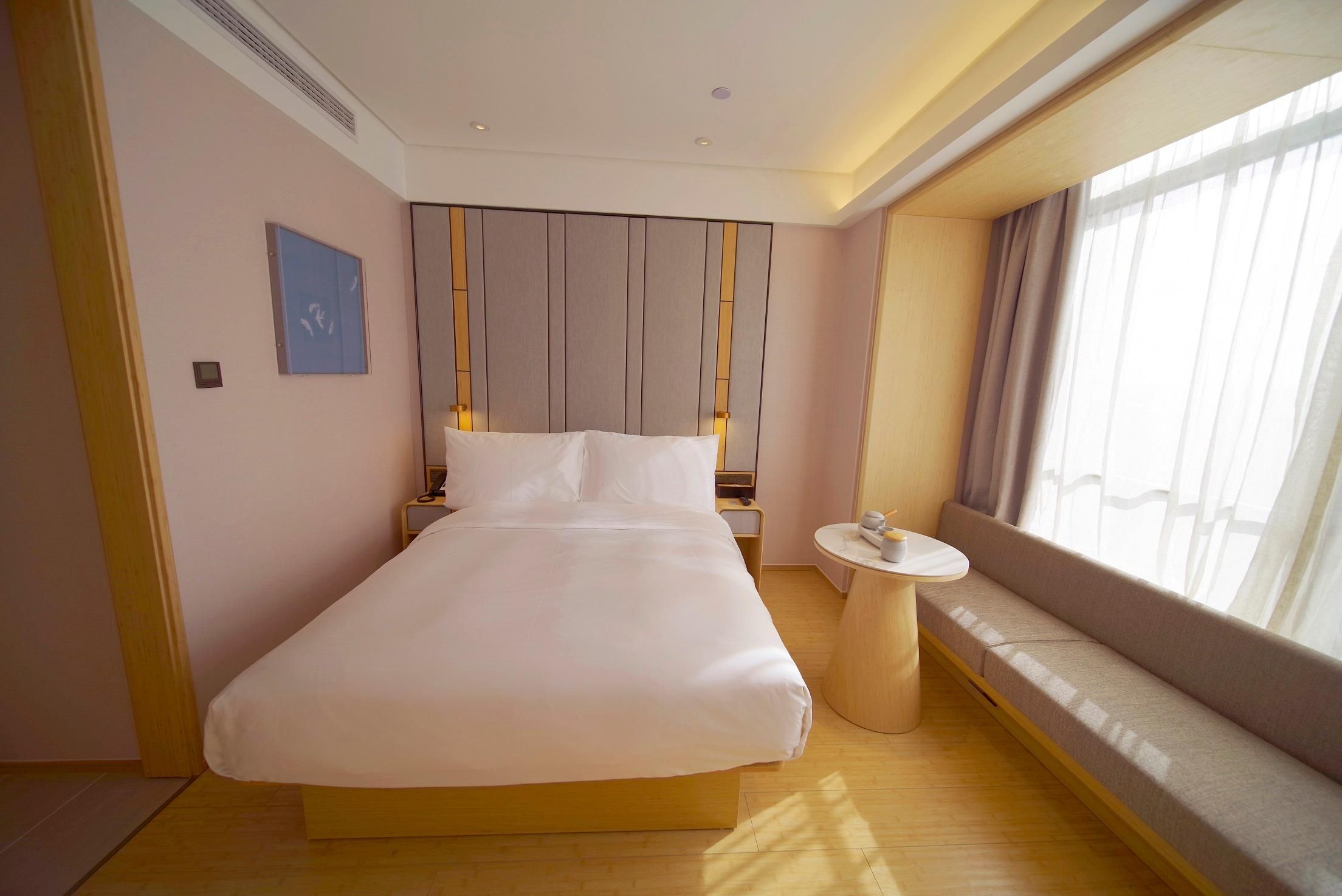 Een hotelkamer met een groot bed, een nachtkastje, een klein rond tafeltje, een bank bij het raam en een paneel aan de muur.