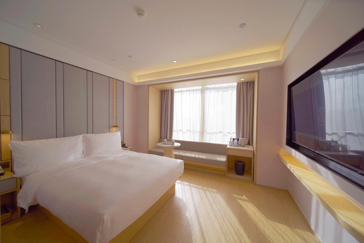 Una habitación de hotel con una cama grande, televisión, ventana con cortinas, una mesita pequeña y un banco.