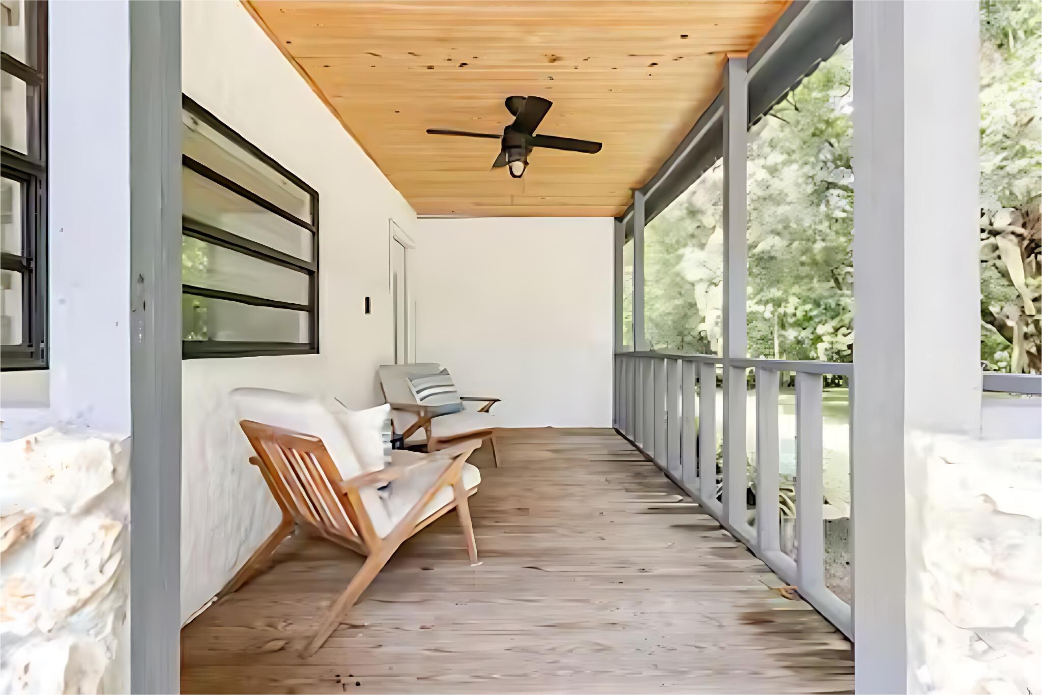 Un porche cubierto con ventilador de techo, dos sillas y vista a árboles.