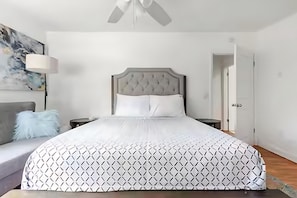 Ein ordentlich bezogenes Bett mit einem geometrisch gemusterten Überwurf, einem gepolsterten Kopfteil und einem grauen Sofa mit einem flauschigen Kissen.