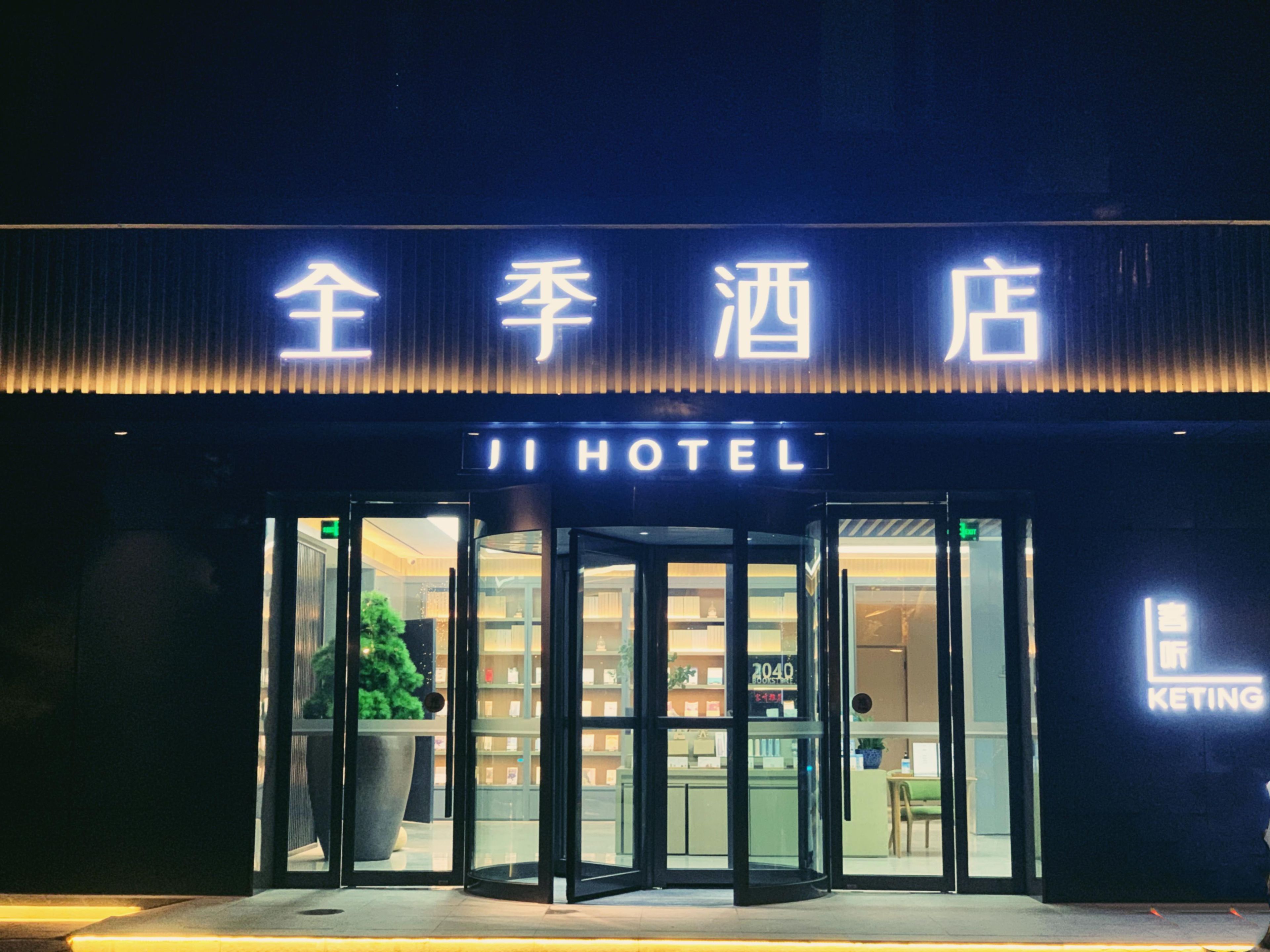 Entrada de un hotel con puerta giratoria, una planta grande en maceta y un letrero con caracteres chinos y la palabra 'HOTEL' en inglés.