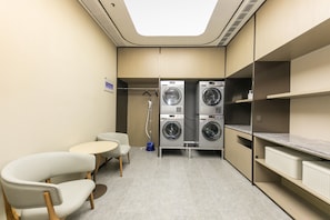 A compact laundry room with two washing machines, a small table, two chairs, and a ceiling light. - Ji Hotel (Nanjing)