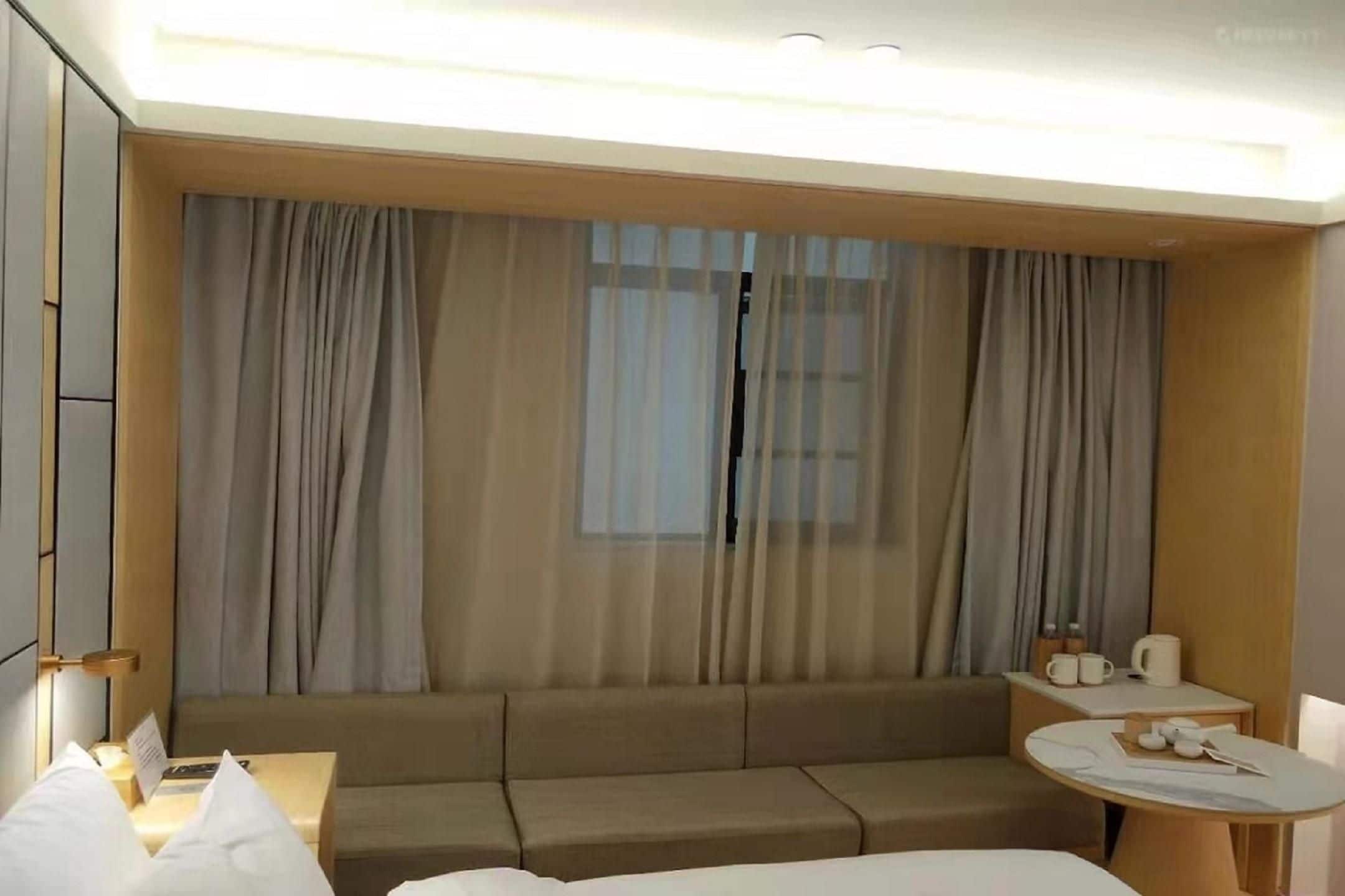 Hotellihuone, jossa on vuodesohva, yöpöytä lampun kanssa, pieni pyöreä pöytä, jossa on kuppi ja aluslautanen, sekä ikkuna, jossa on ohuet verhot.