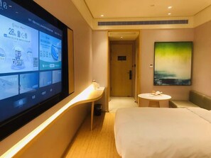A hotel room with a bed, a desk, a TV displaying hotel information, a painting on the wall, and a door leading to another room.