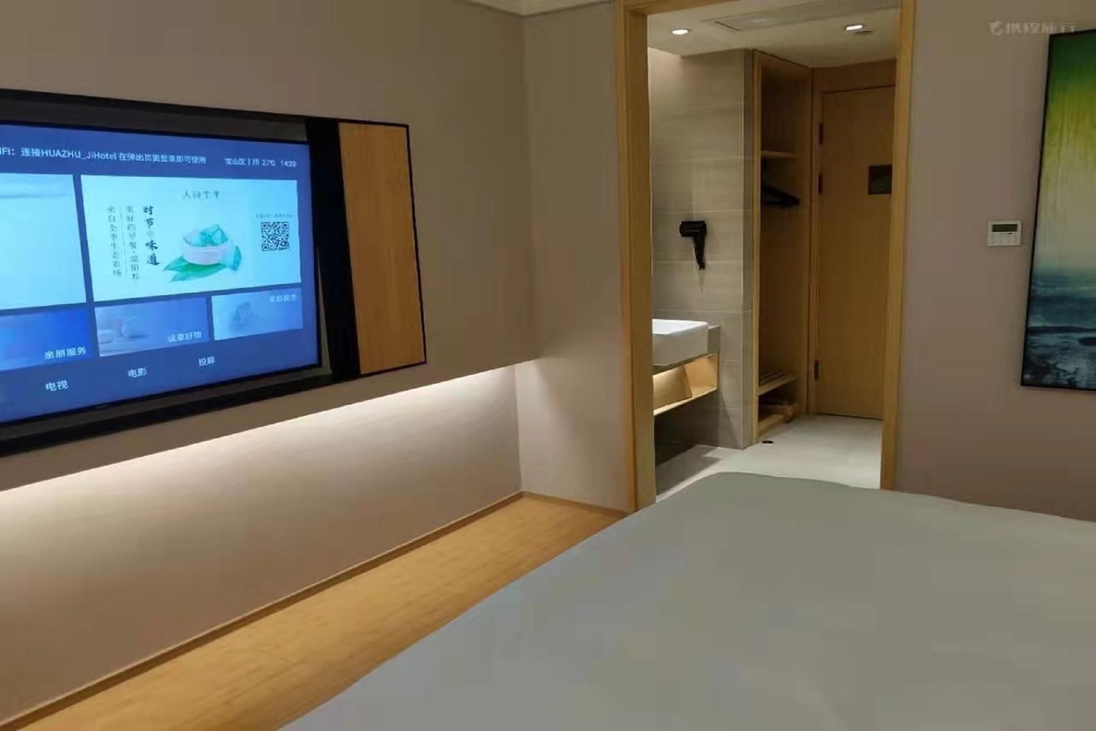 Une chambre d’hôtel dotée d’un grand écran numérique affichant des informations, d’un plancher en bois, d’un lit blanc et d’une porte donnant accès à une salle de bain.