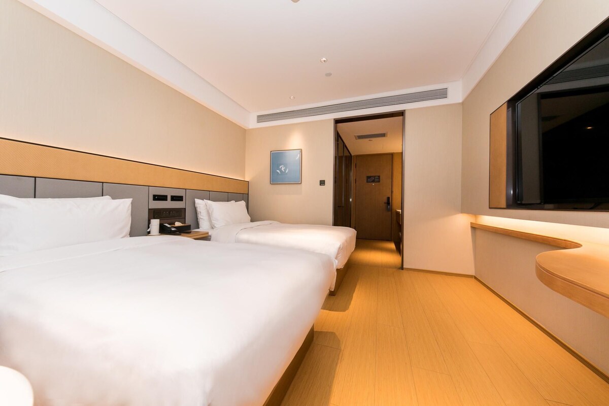 A hotel room with two beds, a bedside table with a phone, a small bedside table with a lamp, a window, and a door leading to another room.