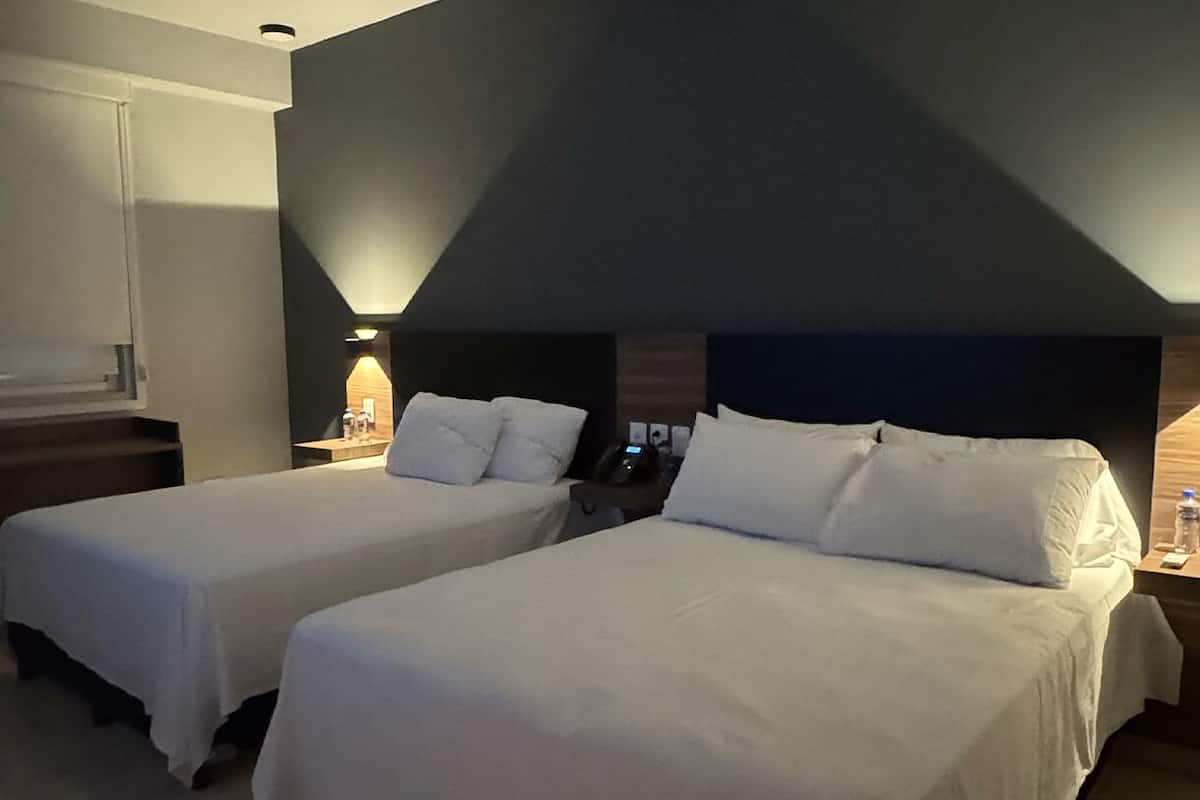 A hotel room with two beds, a bedside table with a lamp, a phone, and a wall-mounted light fixture.