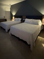 Habitación de hotel con dos camas, una mesita de noche con lámpara, teléfono y luz empotrada en la pared.
