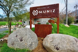 A welcome sign for Muniz Village with a rustic wooden structure, large rocks with handprints, and a grassy landscape. - Sequoia Retreat: BBQ, Patio, Near National Park (Three Rivers)