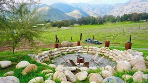 A circular stone fire pit with chairs, surrounded by rocks and grass, set in a grassy field with mountains in the background. - Sequoia Retreat: BBQ, Patio, Near National Park (Three Rivers)