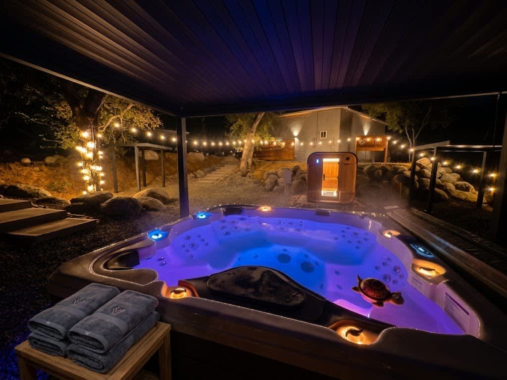 A hot tub with a wooden deck and a covered area, surrounded by string lights and a stone wall.