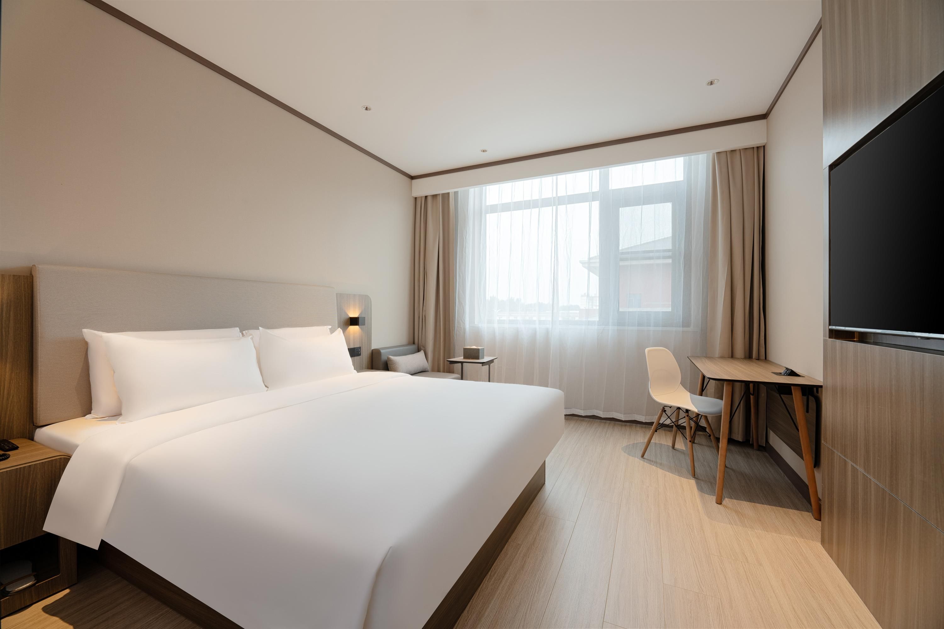 A hotel room with a large bed, a desk, a chair, a TV, and a window with curtains.
