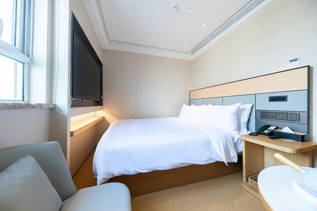 A modern hotel room with a bed, bedside table, TV, and a window with curtains.