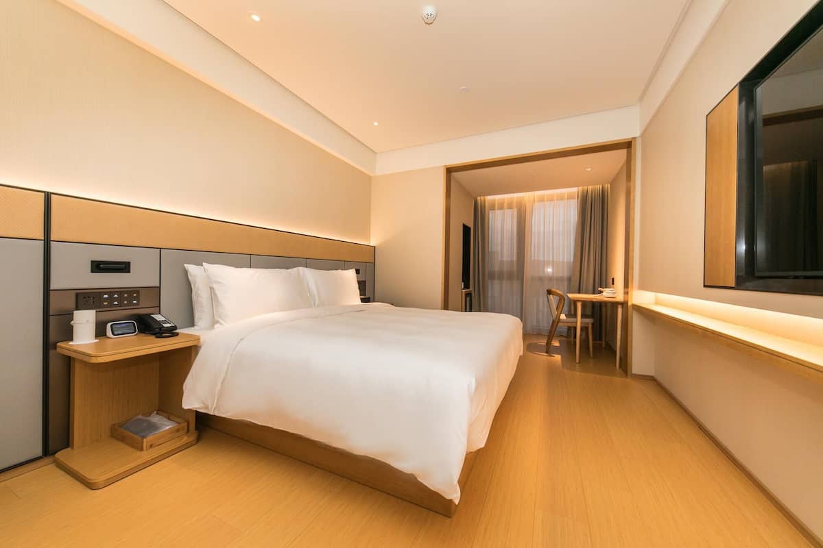 Ett modernt hotellrum med en stor säng, sängbord med telefon och kaffebryggare, ett skrivbord med stol och ett stort fönster med gardiner.