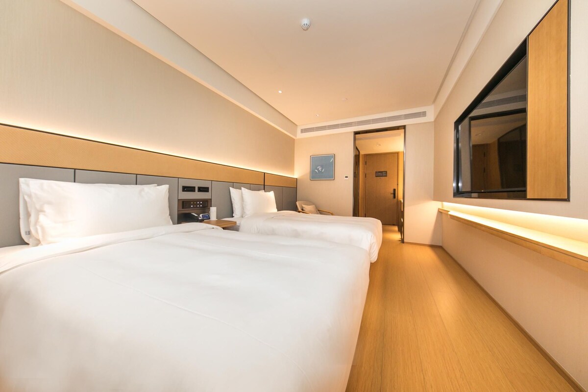 A hotel room with two beds, a bedside table, a wall-mounted TV, and a door leading to another room.