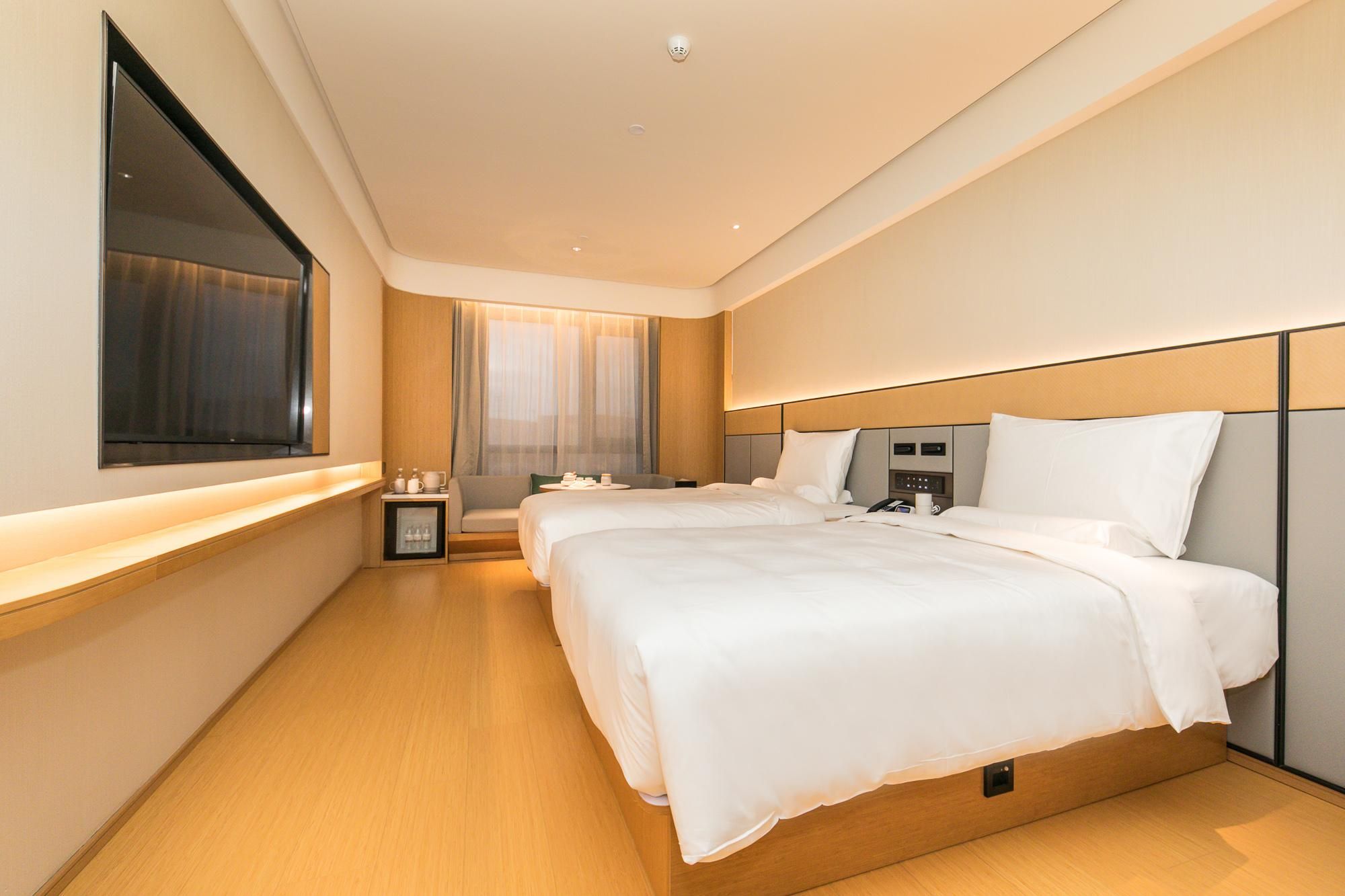 Una habitación de hotel moderna con dos camas, un televisor, una mesita de noche con lámpara y una ventana con cortinas.