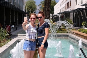 Two individuals taking a selfie in front of a fountain with water jets, surrounded by buildings and greenery. - Kemer Suit (Kemer)