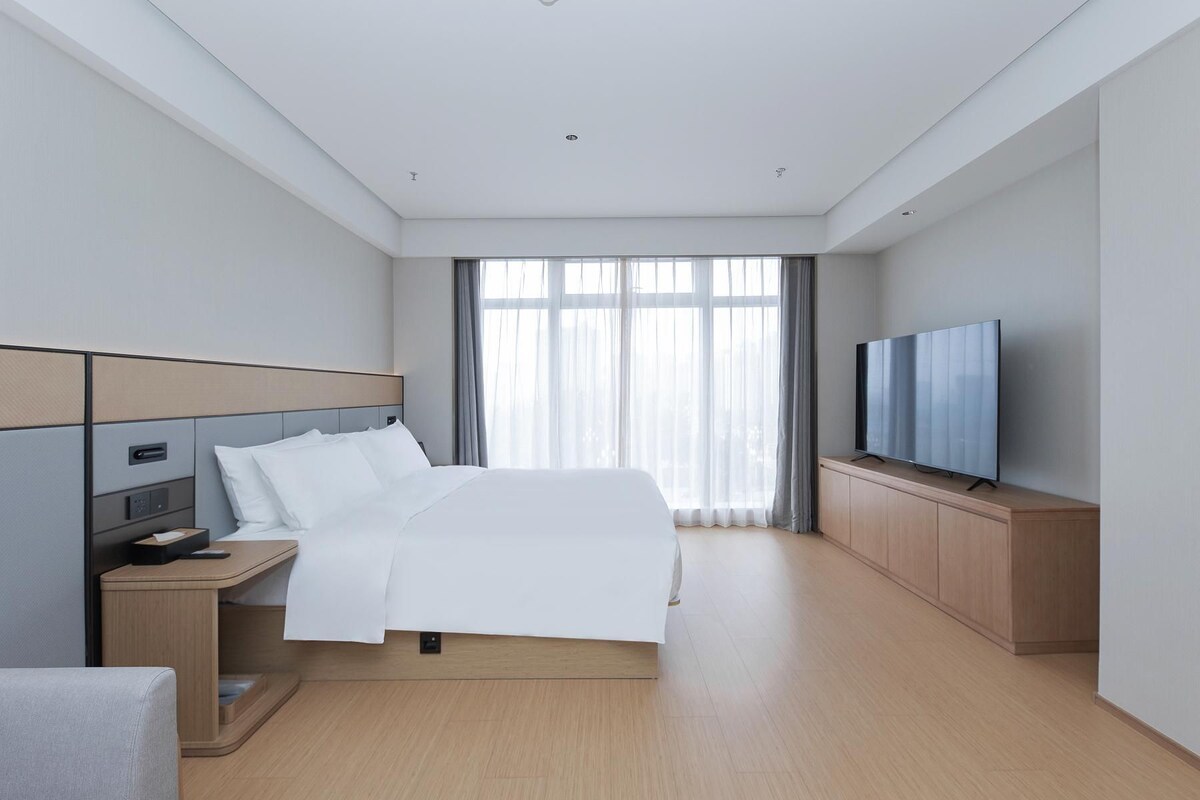 A modern hotel room with a bed, bedside table, TV, and a large window with curtains.