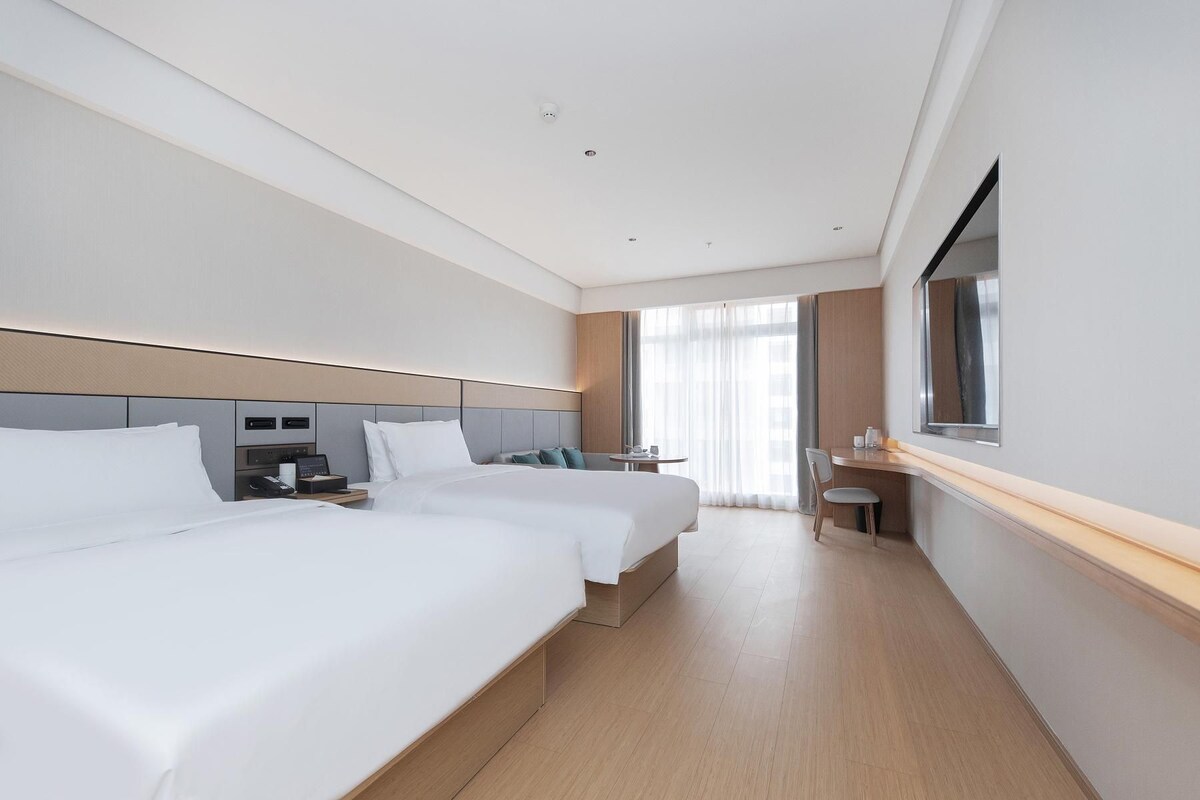 A hotel room with two beds, a desk, a chair, a small table, and a window with curtains.