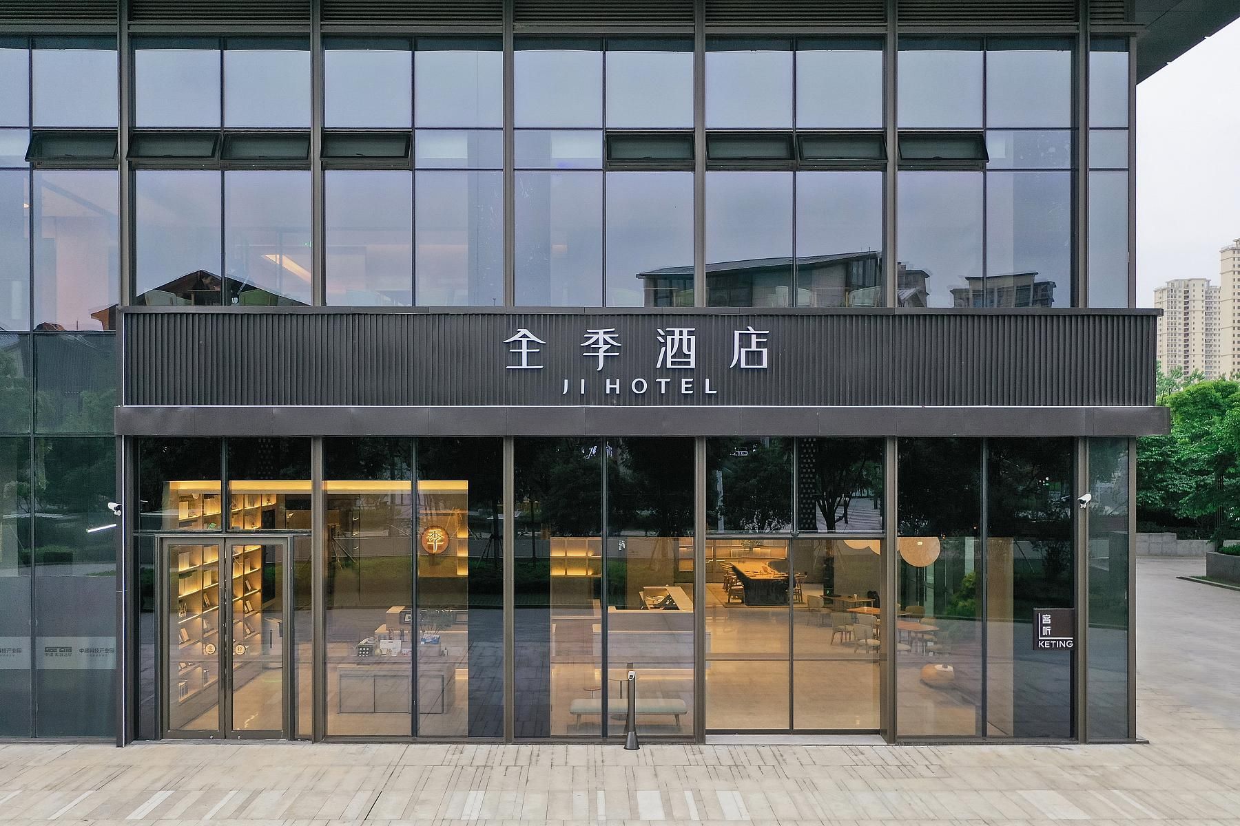 Um hotel moderno com fachada de vidro, área de recepção visível com assentos e uma placa com o nome 'JI HOTEL'.