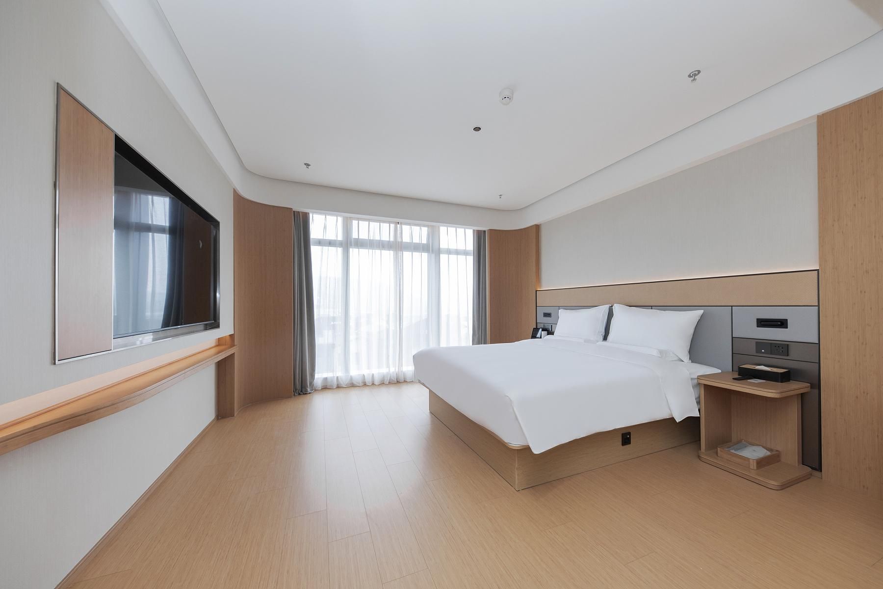 Une chambre d’hôtel moderne dotée d’un grand lit, d’un téléviseur fixé au mur, d’une table de chevet avec une lampe et d’une grande fenêtre avec des rideaux.