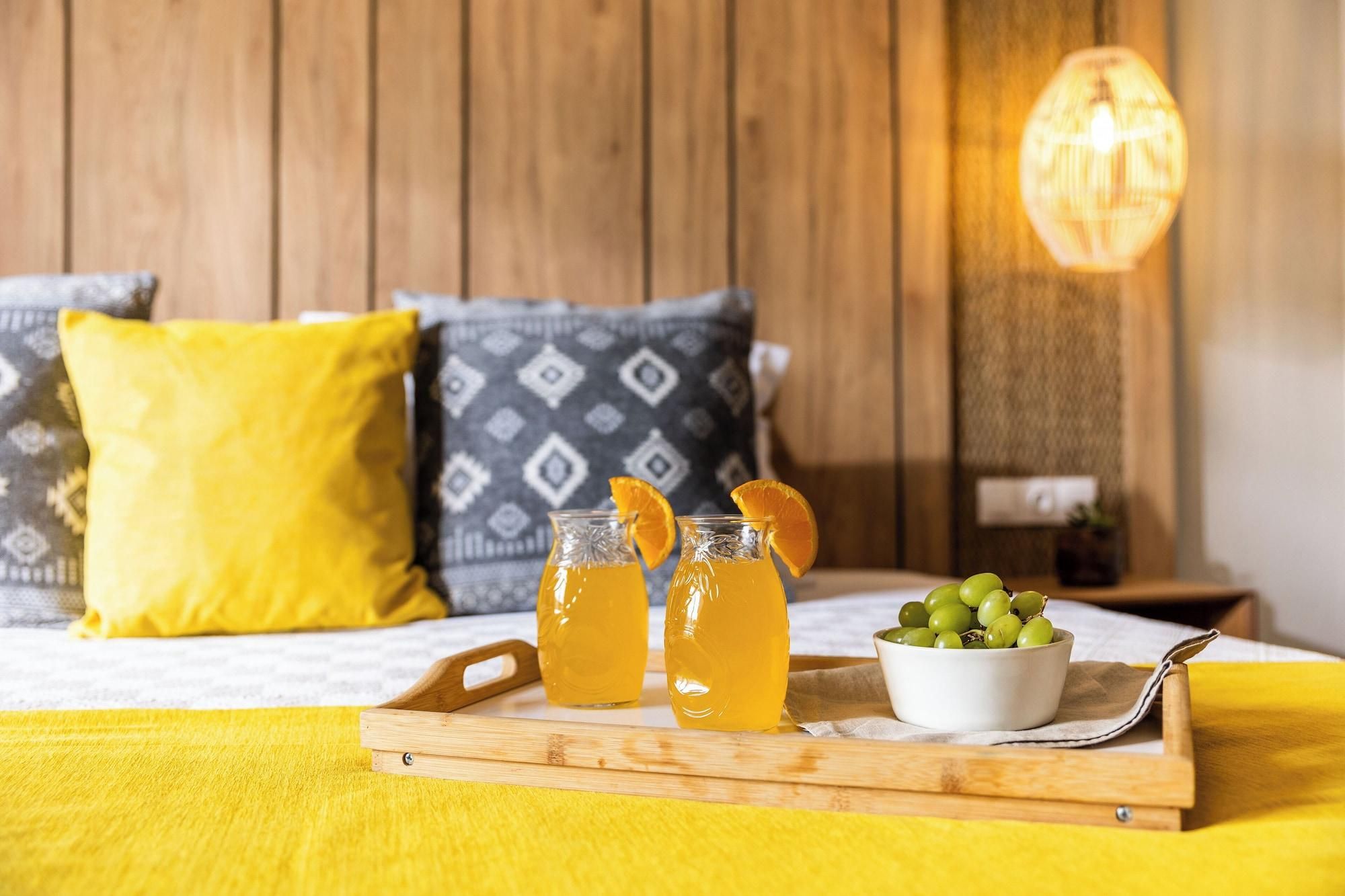 En säng med gula och mönstrade kuddar, ett brickbord med drycker och en skål med vindruvor samt en hängande lampa.