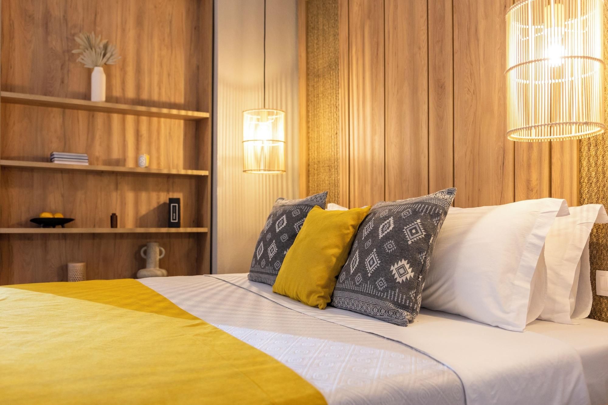 Habitación de hotel con pared de paneles de madera, cama con almohadones estampados, mesita de noche y lámpara colgante.