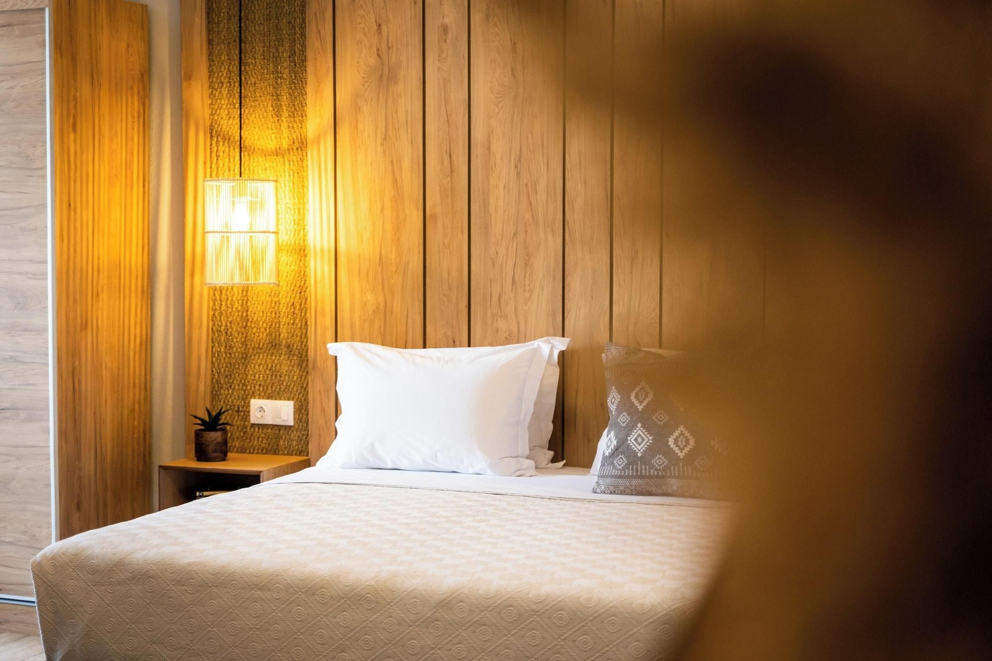 A hotel room with a wooden panel wall, a bed with white linens, a nightstand with a lamp, and a small plant.