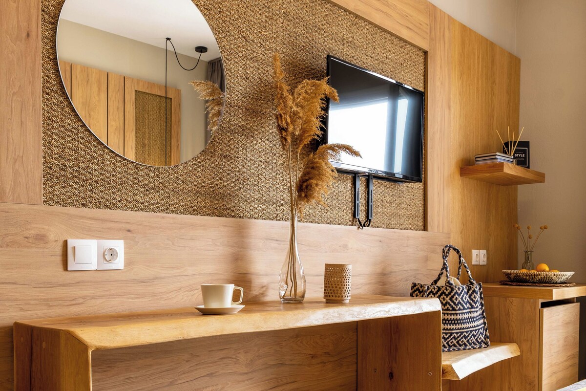 A modern bathroom with a wooden vanity, a round mirror, a television, and a small shelf with decorative items.