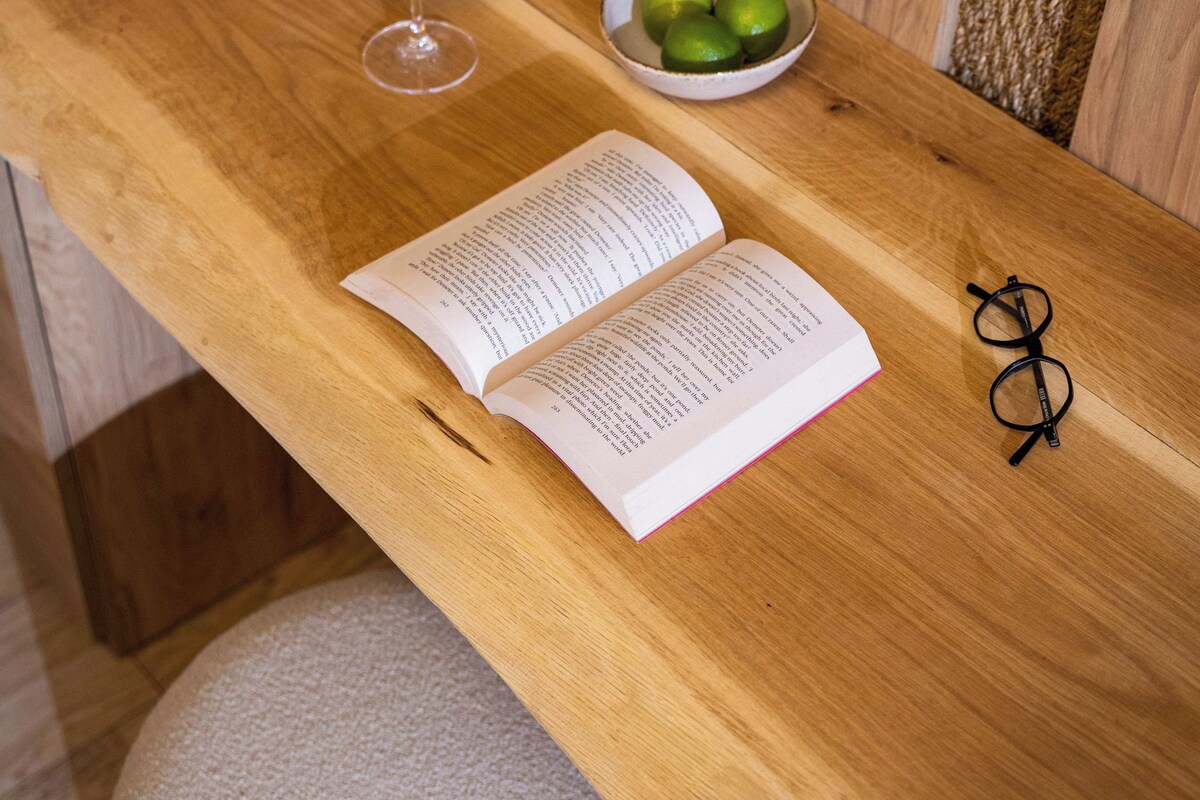 A wooden table with a book, a pair of glasses, a glass with a lime slice, and a bowl of limes.