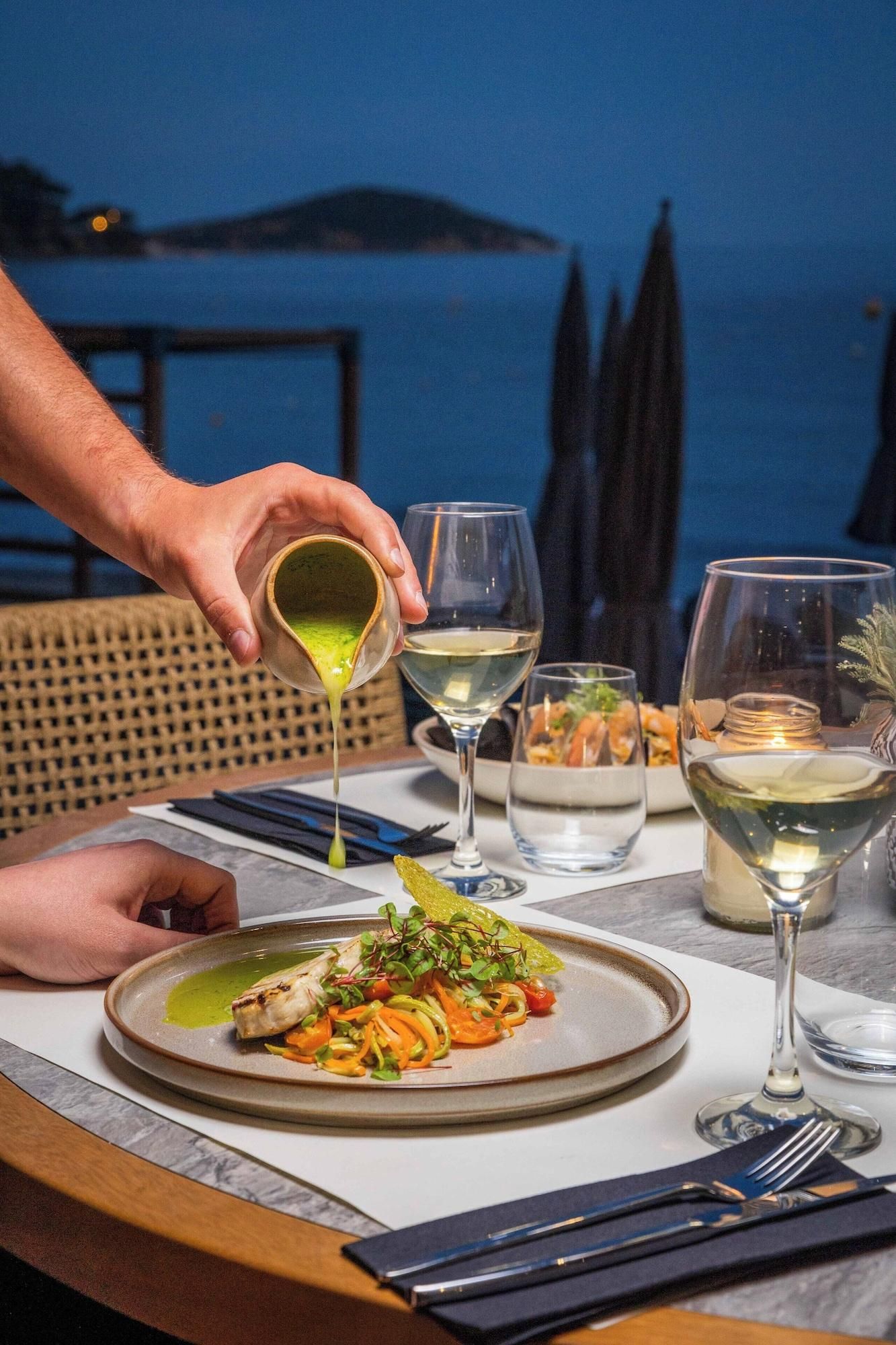 A dining table with a plate of food, two glasses of white wine, and a person pouring a green sauce.