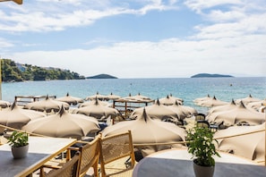 A beachside restaurant with numerous beige umbrellas and wicker furniture, overlooking a calm sea and distant hills. - Skiathos Thalassa (Skiathos)