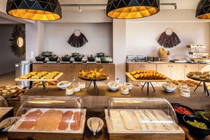 A buffet setup with various food items, including meats and pastries, under modern pendant lighting. - Skiathos Thalassa (Skiathos)