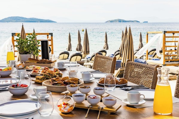 A dining table set with various dishes and drinks, overlooking a body of water with boats in the distance. - Skiathos Thalassa (Skiathos)