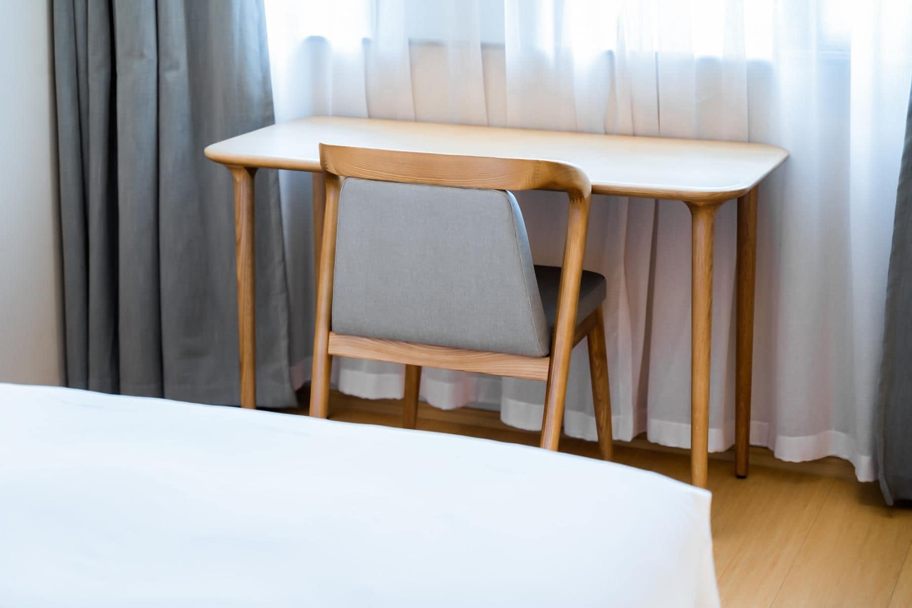 A bed, a wooden chair with a grey cushion, and a wooden desk against a window with curtains.