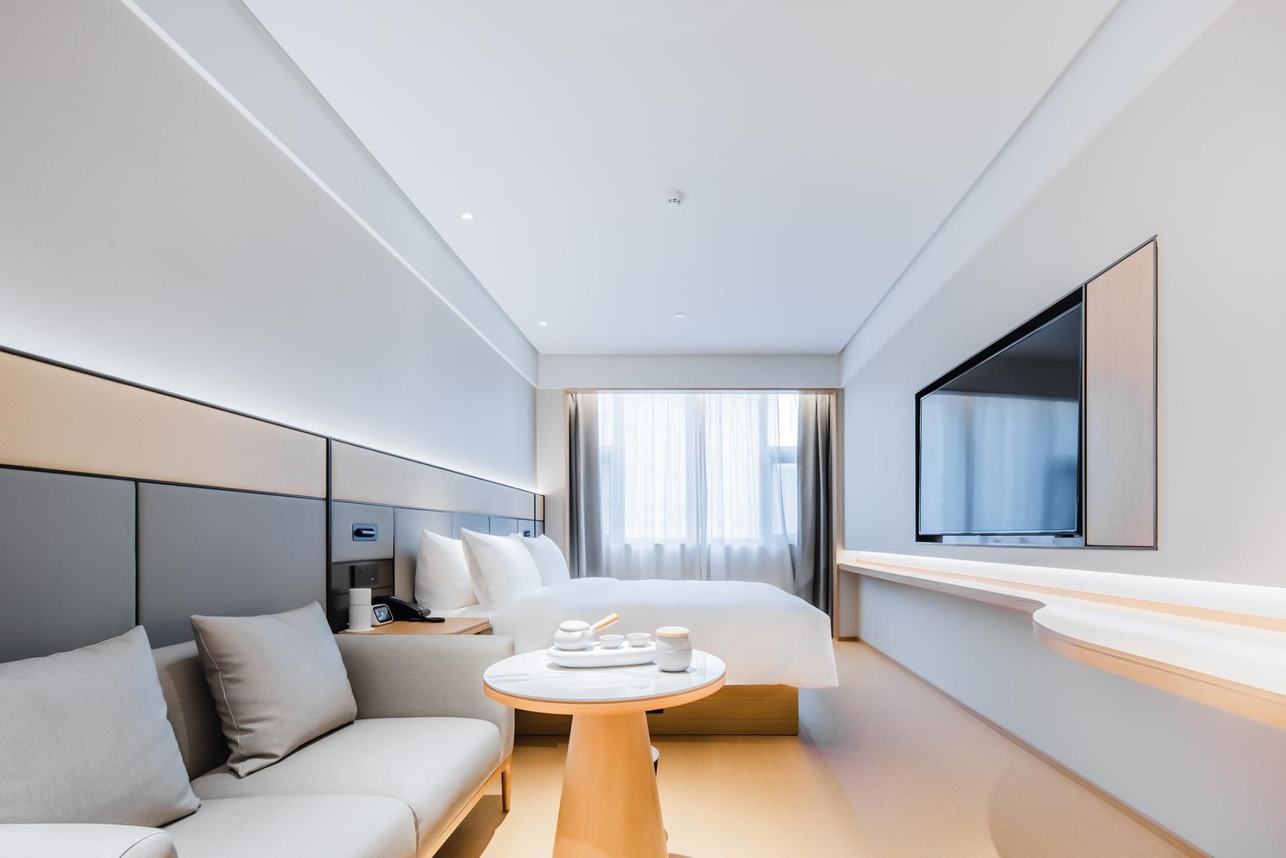 A modern hotel room with a bed, a sofa, a small table with amenities, a TV, and a large window with curtains.