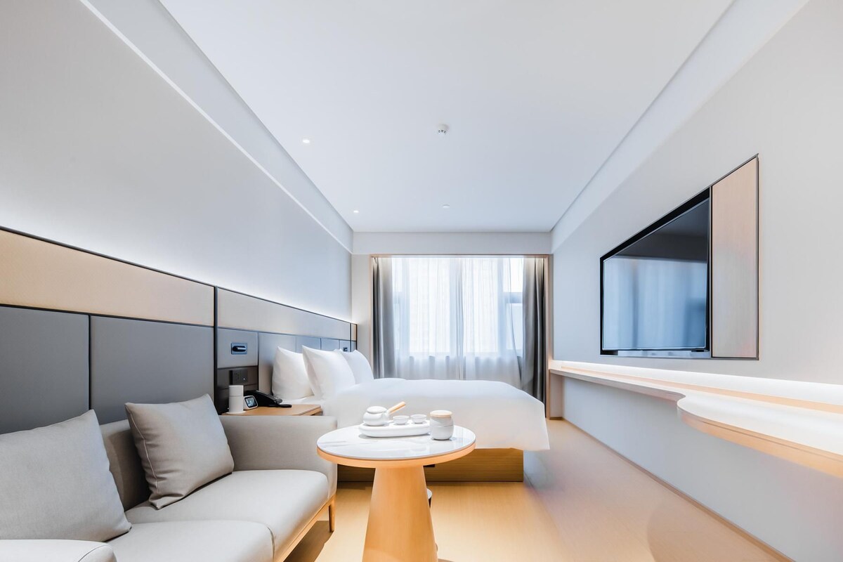 Quarto de hotel moderno com cama, sofá, mesinha com amenidades, televisão e uma grande janela com cortinas.