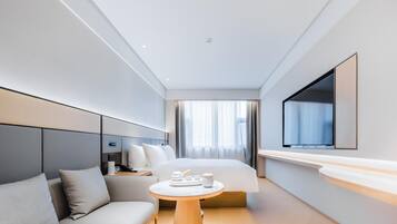 A modern hotel room with a bed, a sofa, a small table with amenities, a TV, and a large window with curtains.