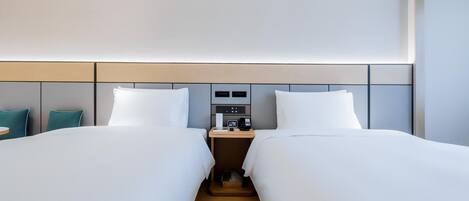 A hotel room with two beds, a bedside table with a lamp, a small table with a phone and a clock, and a wall-mounted control panel.