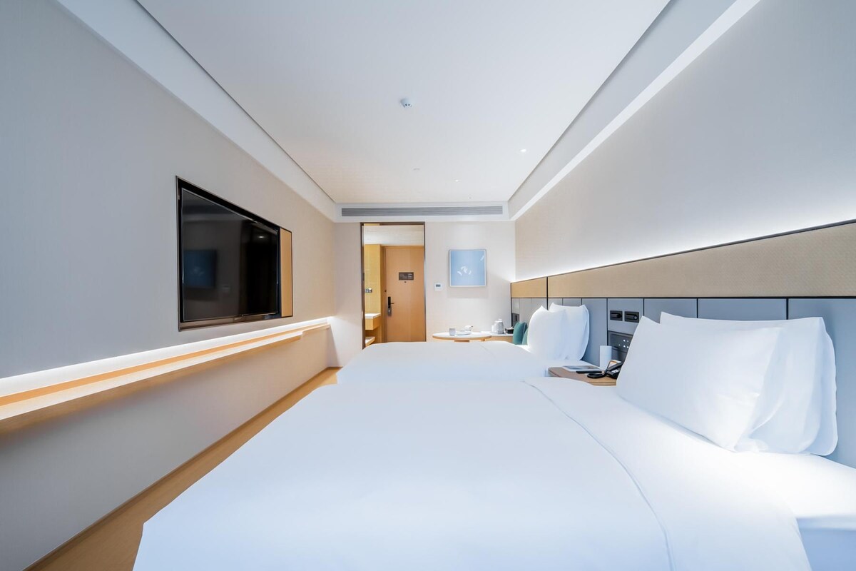 Quarto de hotel moderno com uma cama grande, televisão de tela plana na parede, mesa de cabeceira com abajur e cabeceira com controles integrados.
