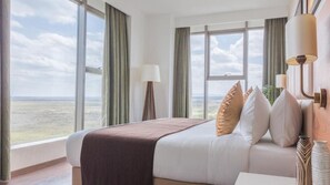 A hotel room with a large bed, two bedside lamps, a view of the landscape, and a window with curtains. - Emara Ole Sereni (Syokimau)