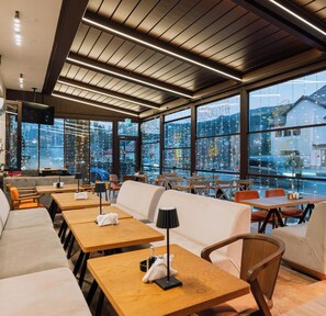 Un restaurante moderno con mesas de madera, sofás blancos y amplios ventanales con vistas a la ciudad.