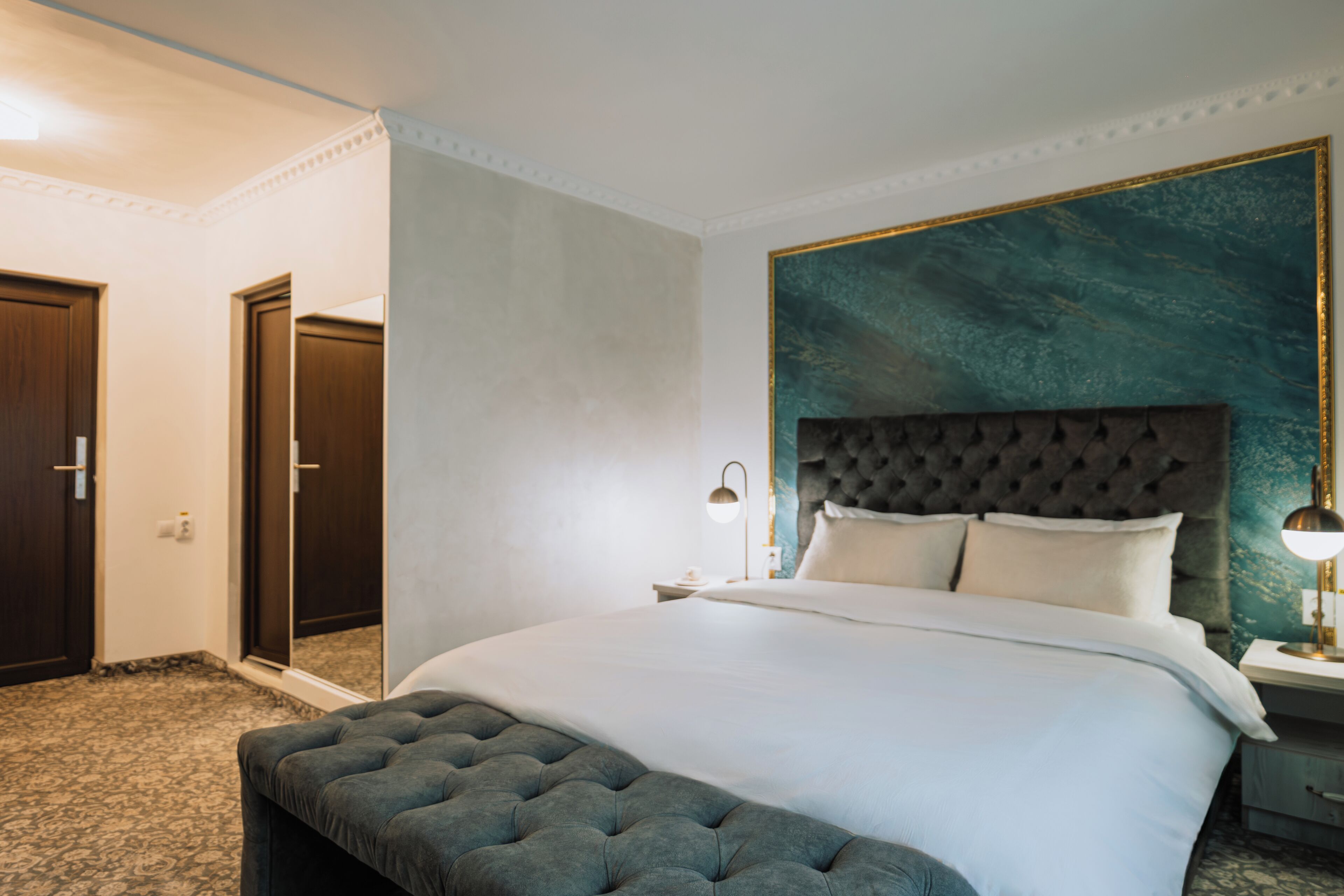 Una habitación de hotel con una cama grande, un cabecero capitonado, un banco al pie de la cama, una mesita de noche con lámpara y un espejo en la pared.
