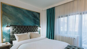 Quarto de hotel com uma cama grande, cabeceira estofada, mesa de cabeceira com abajur e uma grande pintura abstrata na parede.