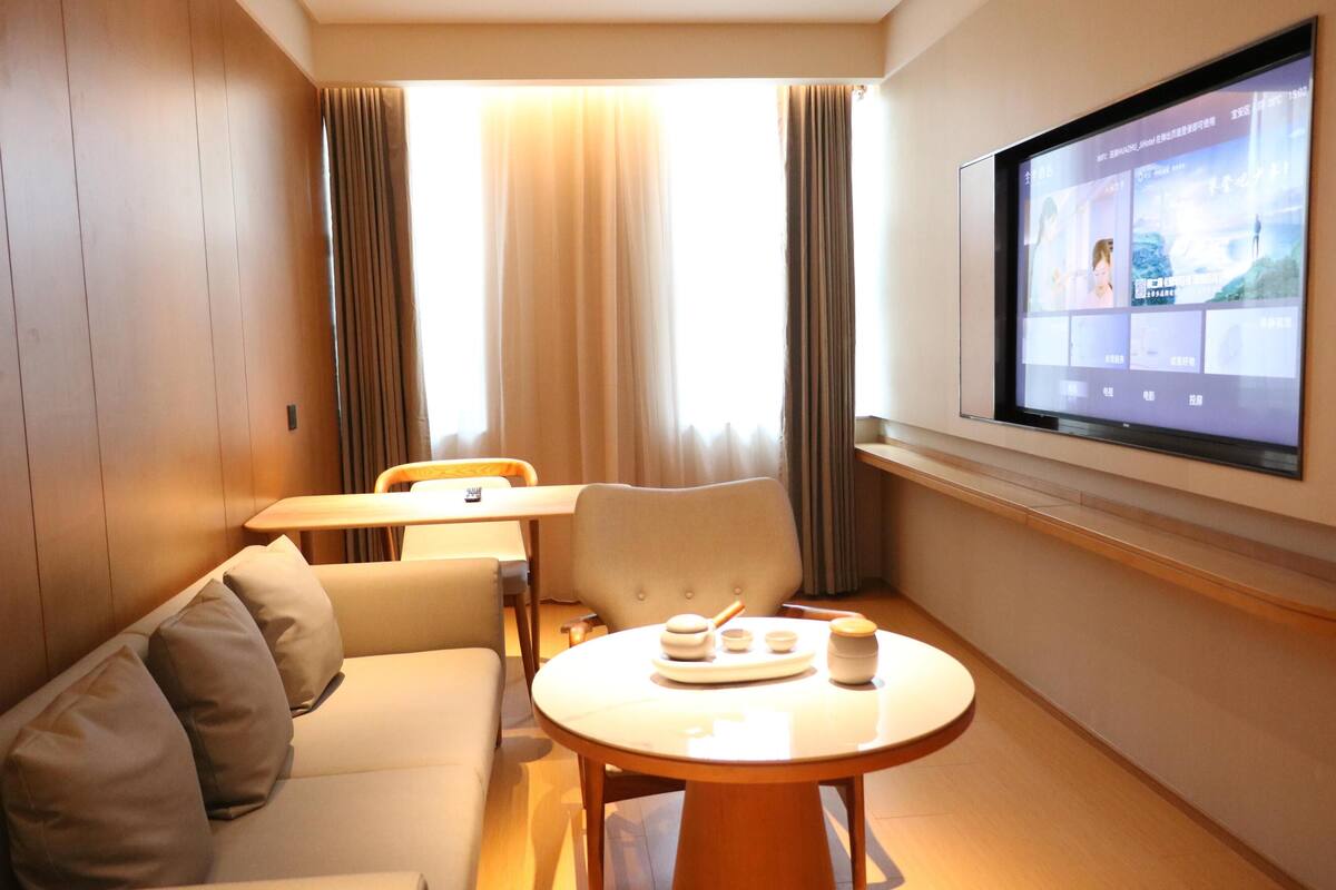 A hotel room with a sofa, a dining table, a TV screen, and a window with curtains.