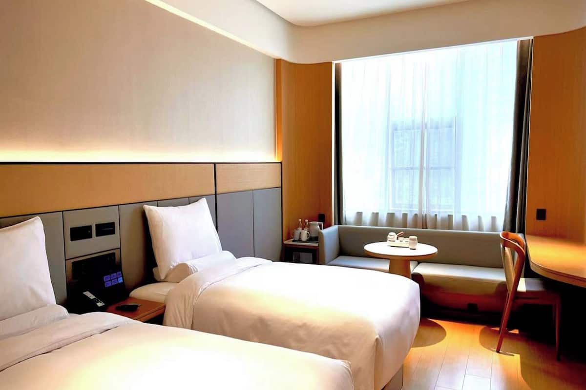 A hotel room with two beds, a small table, a sofa, and a window with curtains.
