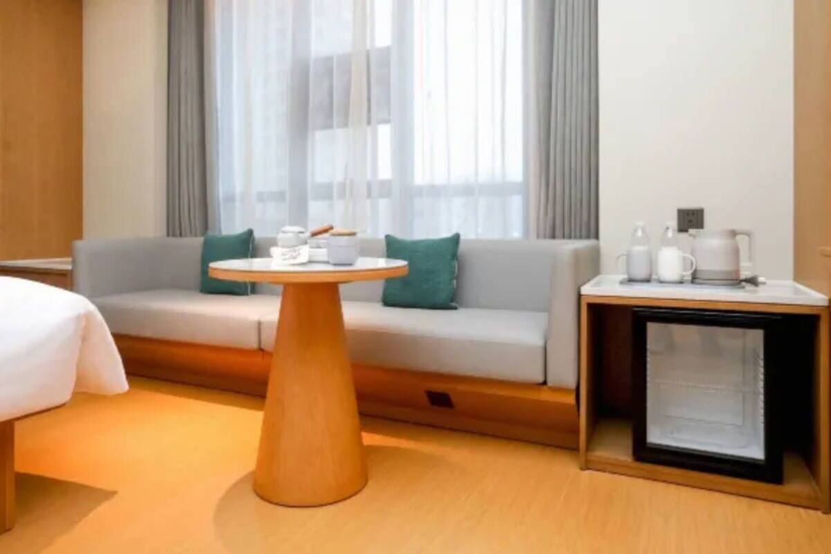 A hotel room with a sofa, a round wooden table, a bedside table with a lamp, and a window with curtains.