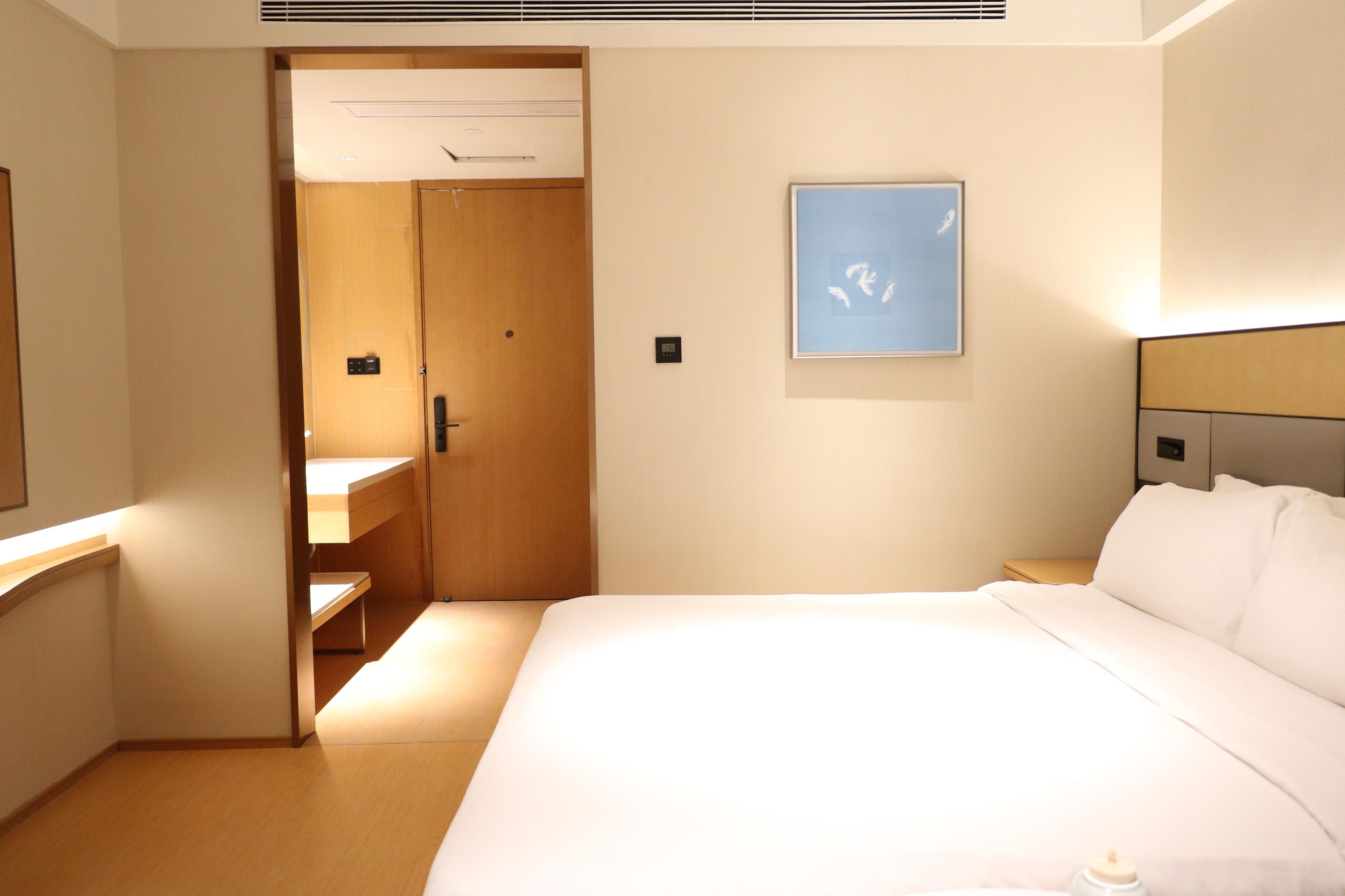 A hotel room with a bed, a bedside table, a mirror, a door leading to a bathroom, and a framed picture on the wall.