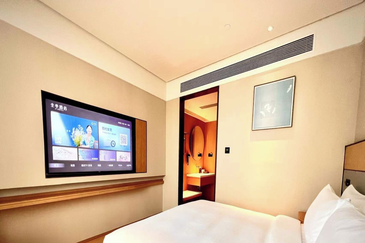 A hotel room with a bed, a TV mounted on the wall, a small table, and a bathroom visible through an open door.