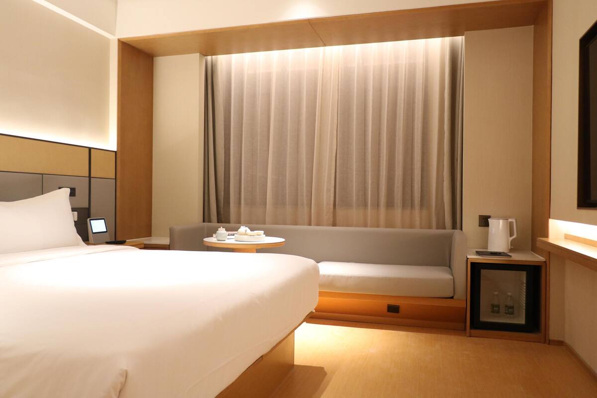 A hotel room with a large bed, a sofa, a small table with a pitcher, and a nightstand with a lamp.